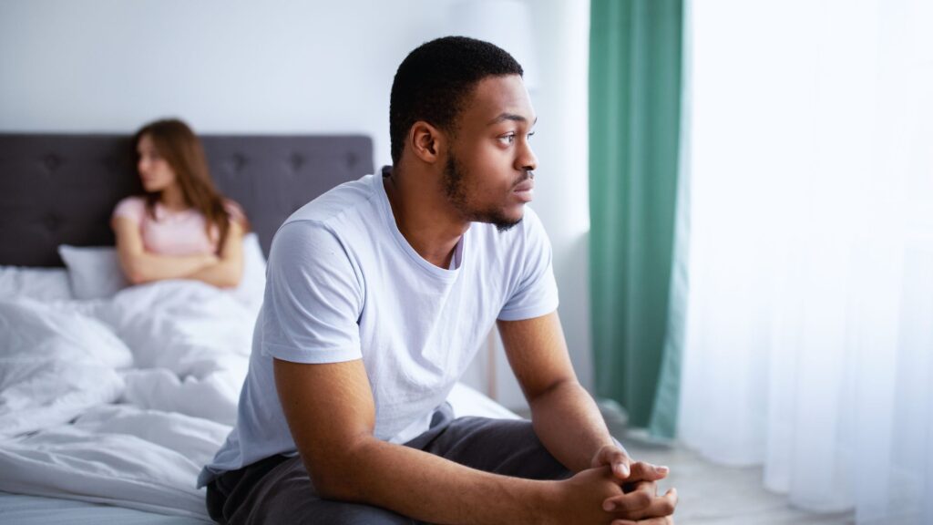 A man sits thoughtfully on a bed, gazing out a window, while a woman with arms crossed sits blurred in the background. Relationship tension.